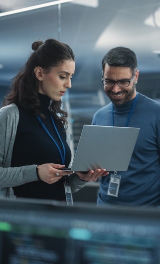 Zwei IT-Mitarbeiter, eine Frau und ein Mann, überprüfen gemeinsam Daten auf einem Laptop im Serverraum. Beide tragen Ausweise und scheinen in ein technisches Gespräch vertieft zu sein.